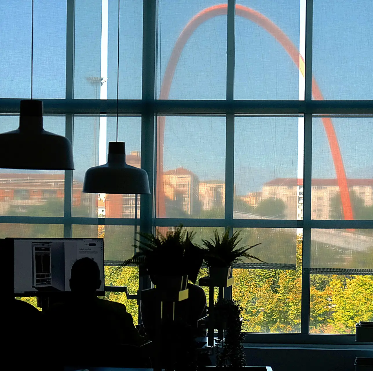 lingotto-camplus Vista dall’interno di ufficio Lingotto Torino con lampade sospese, silhouette lavoratori e Arco Olimpico arancione fuori dalla vetrata