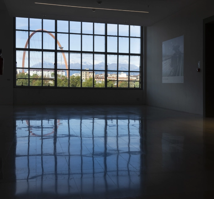 Vista dall’interno del Lingotto Torino attraverso un’ampia vetrata, con riflesso a terra e Arco Olimpico arancione sullo sfondo.