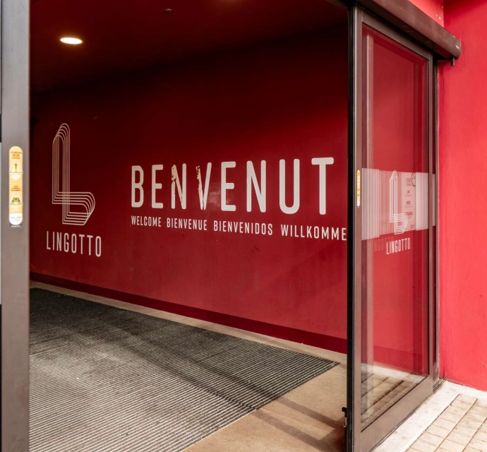 Ingresso rosso Lingotto Torino con porta scorrevole in vetro e scritta “Benvenuto” in varie lingue sulla parete.
