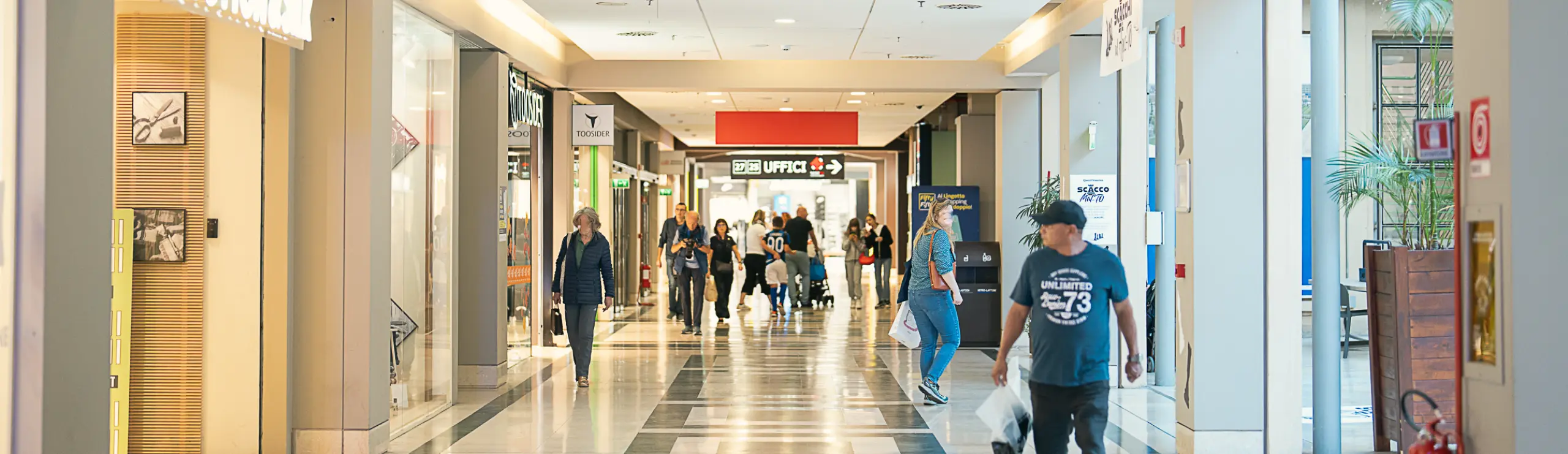 Corridoio interno di centro commerciale con persone che passeggiano e fanno shopping tra i negozi.