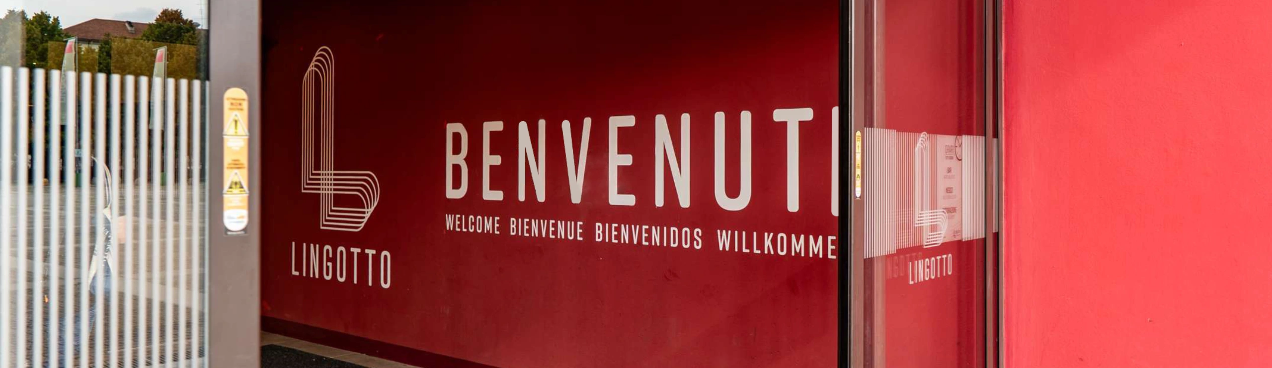 Parete rossa con logo Lingotto Torino, scritta “Benvenuto” in varie lingue all’ingresso del centro commerciale.